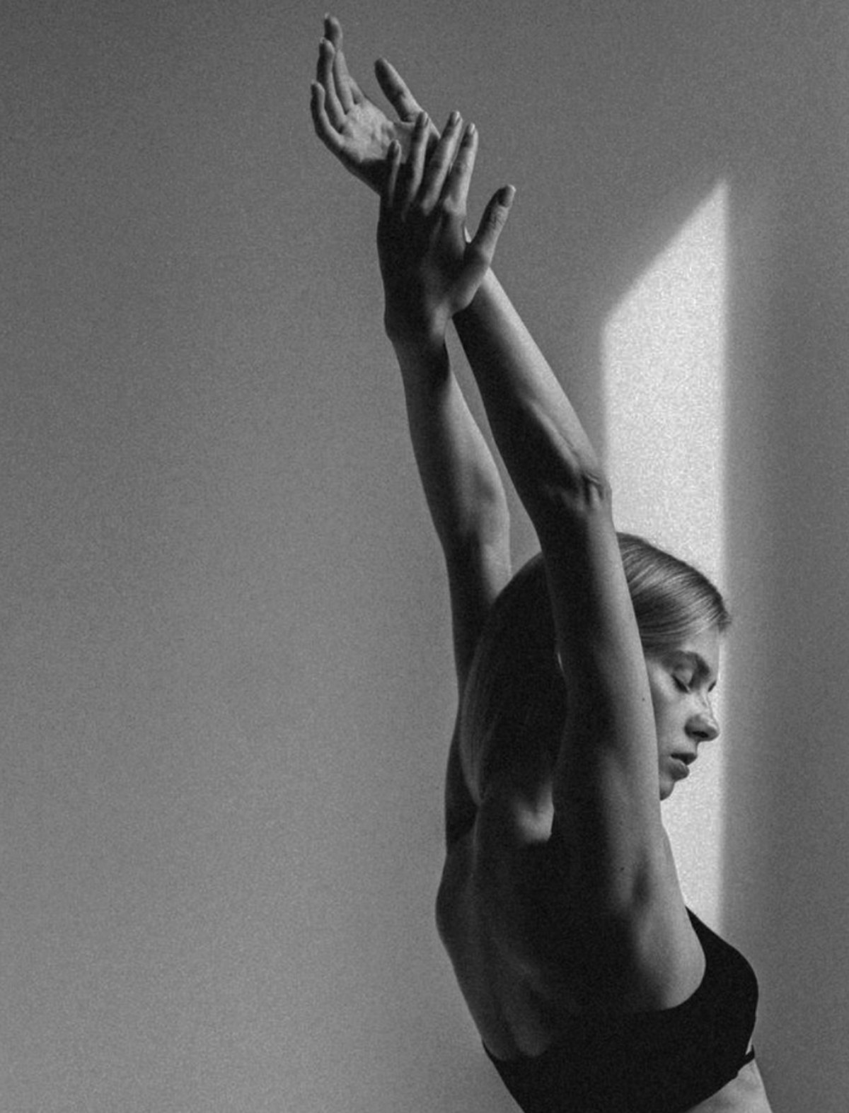 Vrouw in een yogahouding met de armen omhoog, in strijklicht tegen een lichte muur